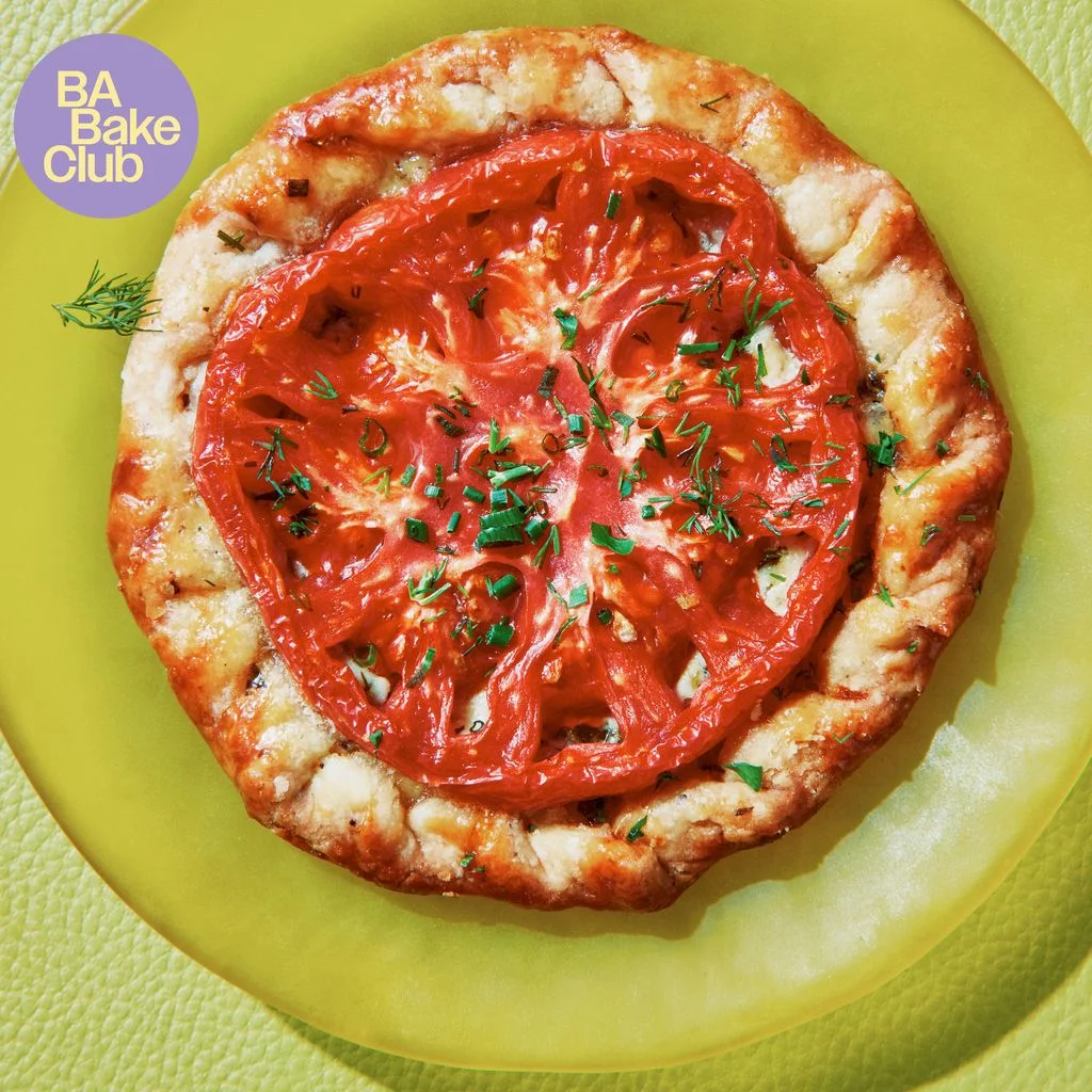 A round of flaky pastry topped with a sliced heirloom tomato chives and dill on a plate.