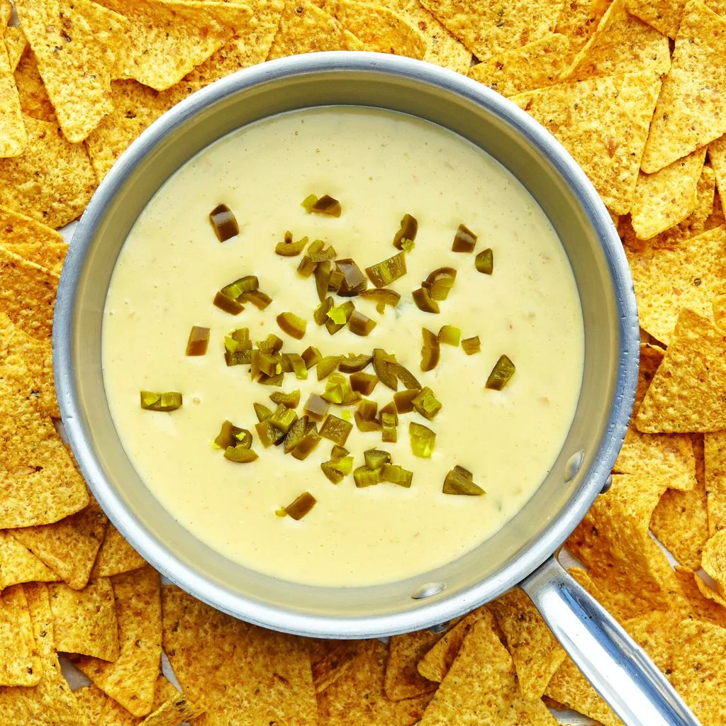 A pot of jalapeñotopped queso dip set in the center of a sheet pan loaded with tortilla chips.