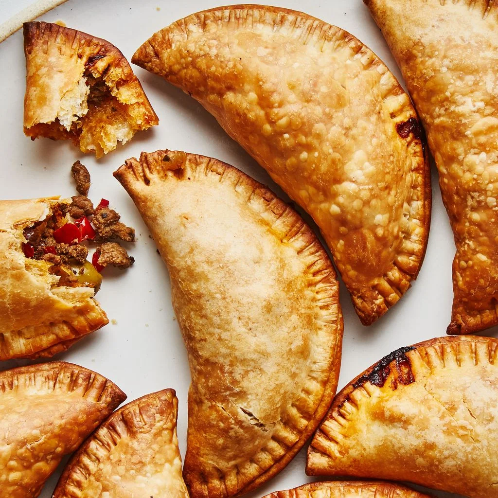 A bunch of forkcrimped beef empanadas on a platter one broken in half to reveal the filling.
