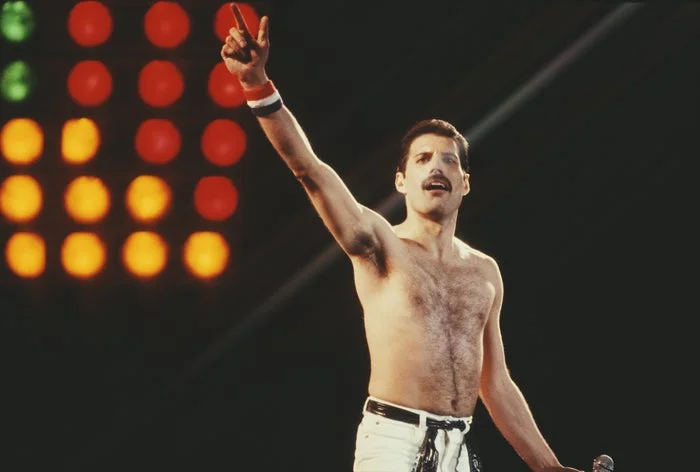 Performer on stage, shirtless with white pants, arm raised holding a microphone stand, lights in the background