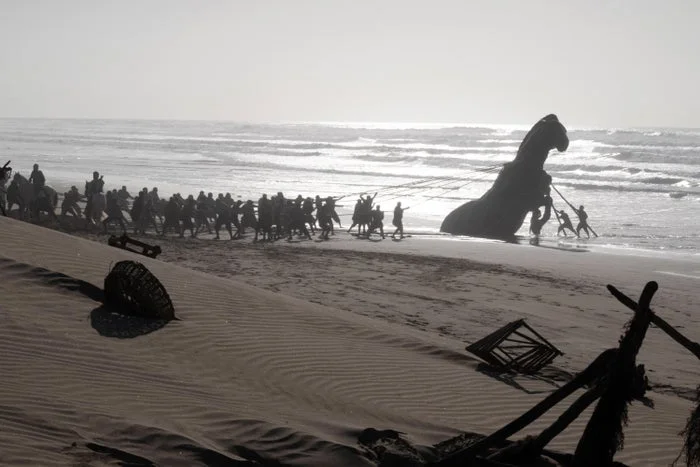 A large group of people pulling a massive ship's hull onto a beach, creating an epic and cinematic scene