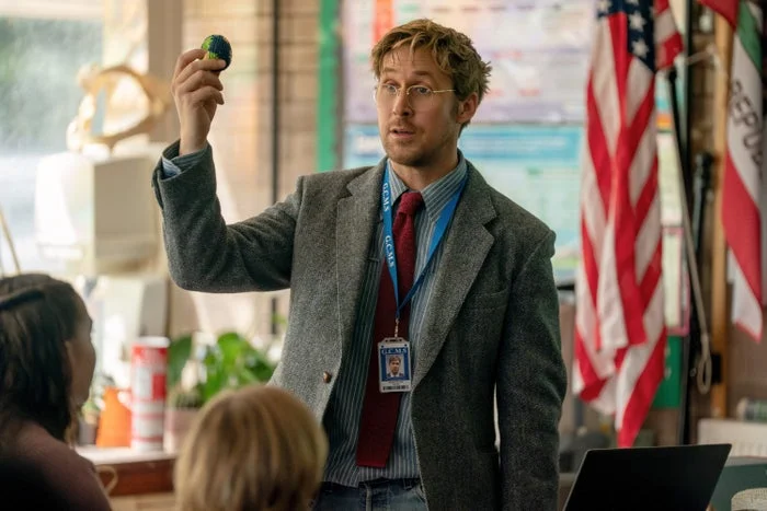 A person in a classroom setting holds up a small globe, dressed in a casual suit with a tie and ID badge. US and California flags in the background