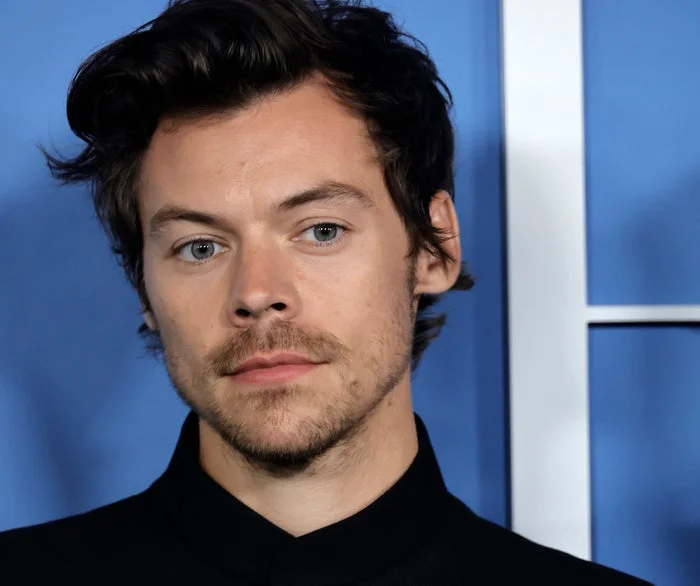 Harry Styles with tousled hair in a jacket stands against a blue background, looking contemplative