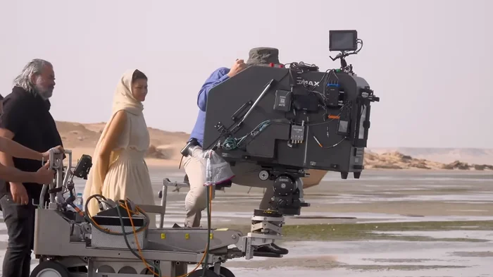 Film crew working in a desert setting with a camera on a dolly; a person in a headscarf and dress stands by