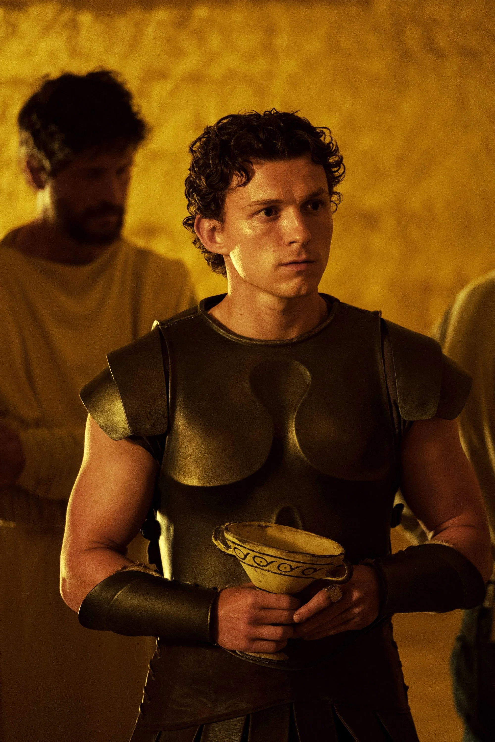 A person in Roman-style armor holds a pottery cup. Another person stands in the background wearing a tunic