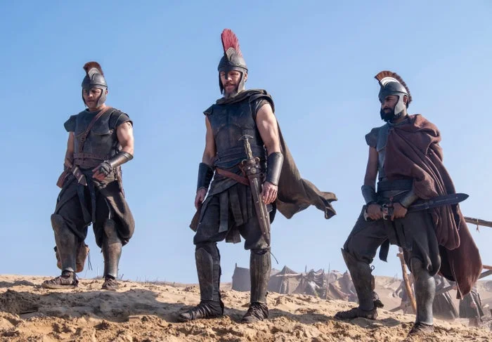 Three people in ancient warrior costumes stand on a sandy landscape, wearing helmets and armor, appearing ready for battle