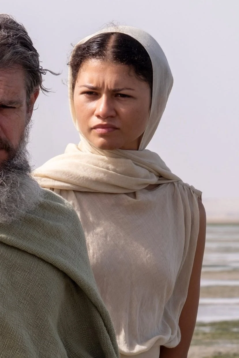 Two people stand near a watery landscape; one wears a green cloak, and the other has a beige headscarf, evoking a historical setting
