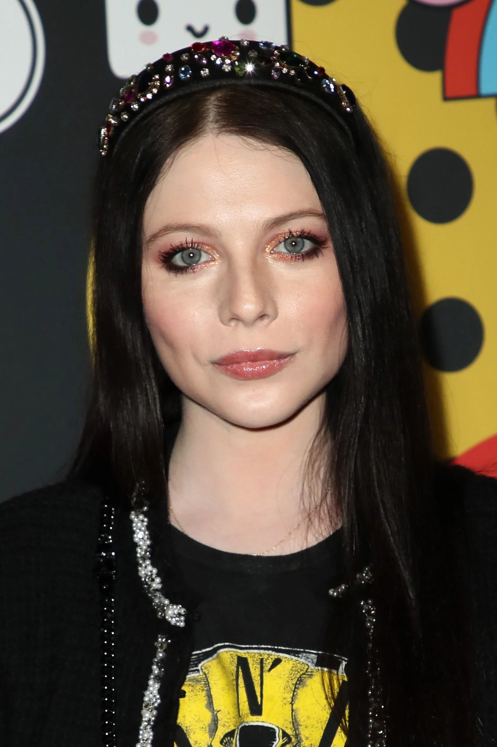 Michelle Trachtenberg with long hair wears an embellished headband and a graphic t-shirt at an event with an artsy backdrop
