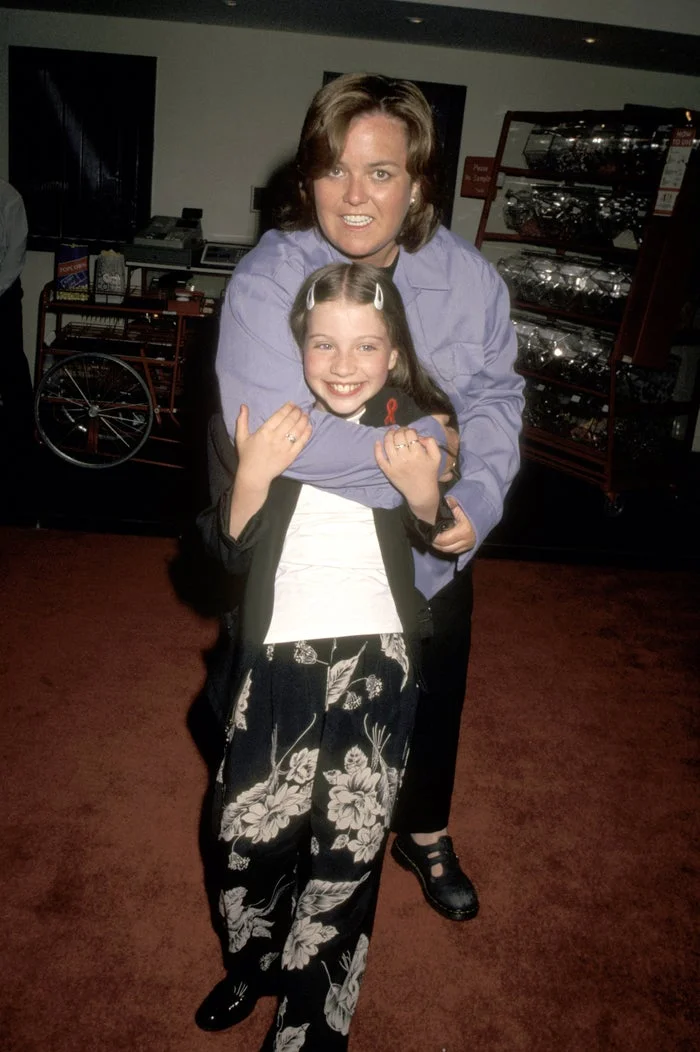 Rosie O'Donnell in a shirt and pants hugs a smiling young Michelle Trachtenberg with floral pants and a jacket