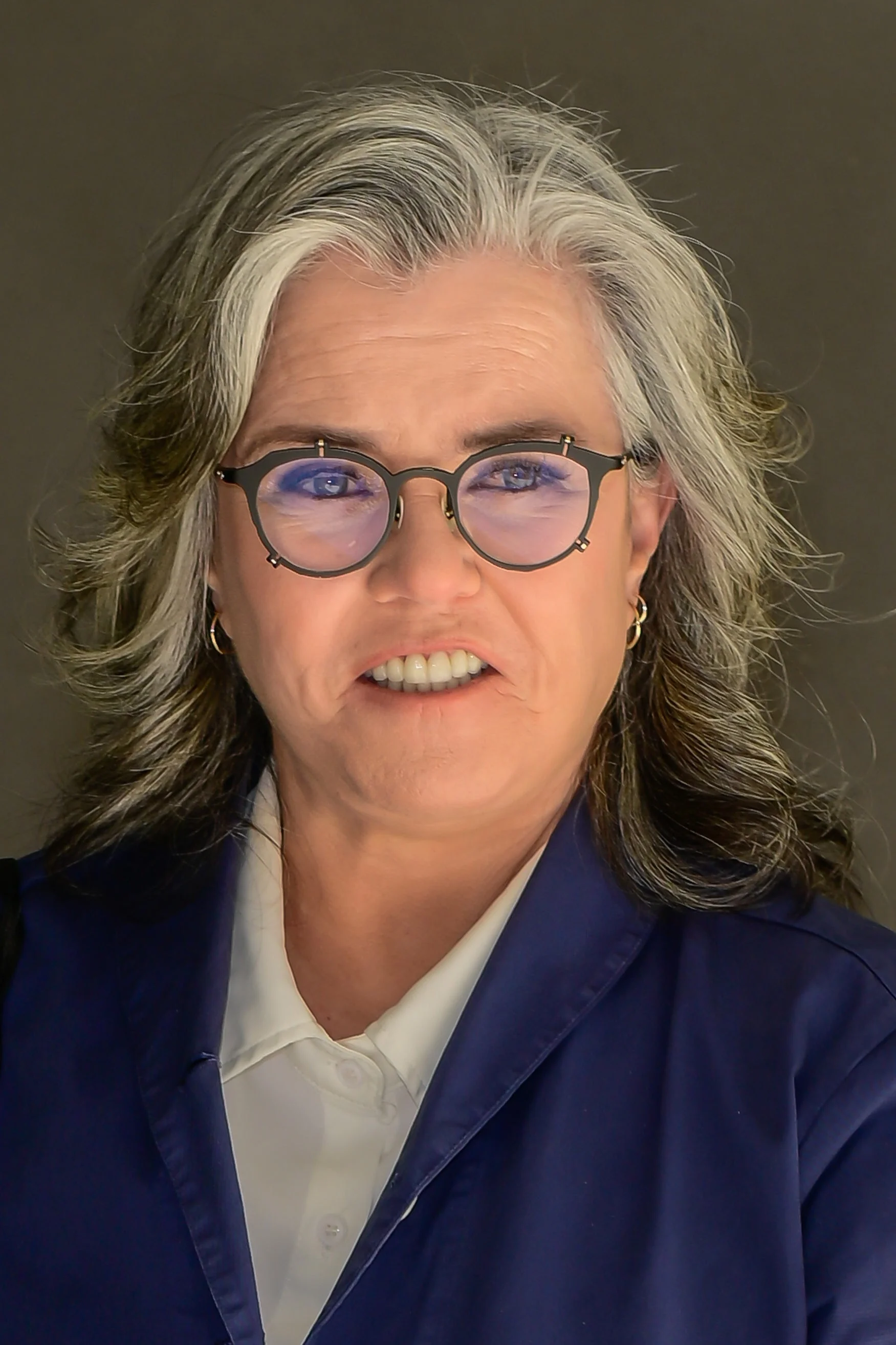 Rosie O'Donnell with glasses and shoulder-length hair, wearing a collared shirt and jacket, smiling slightly at the camera