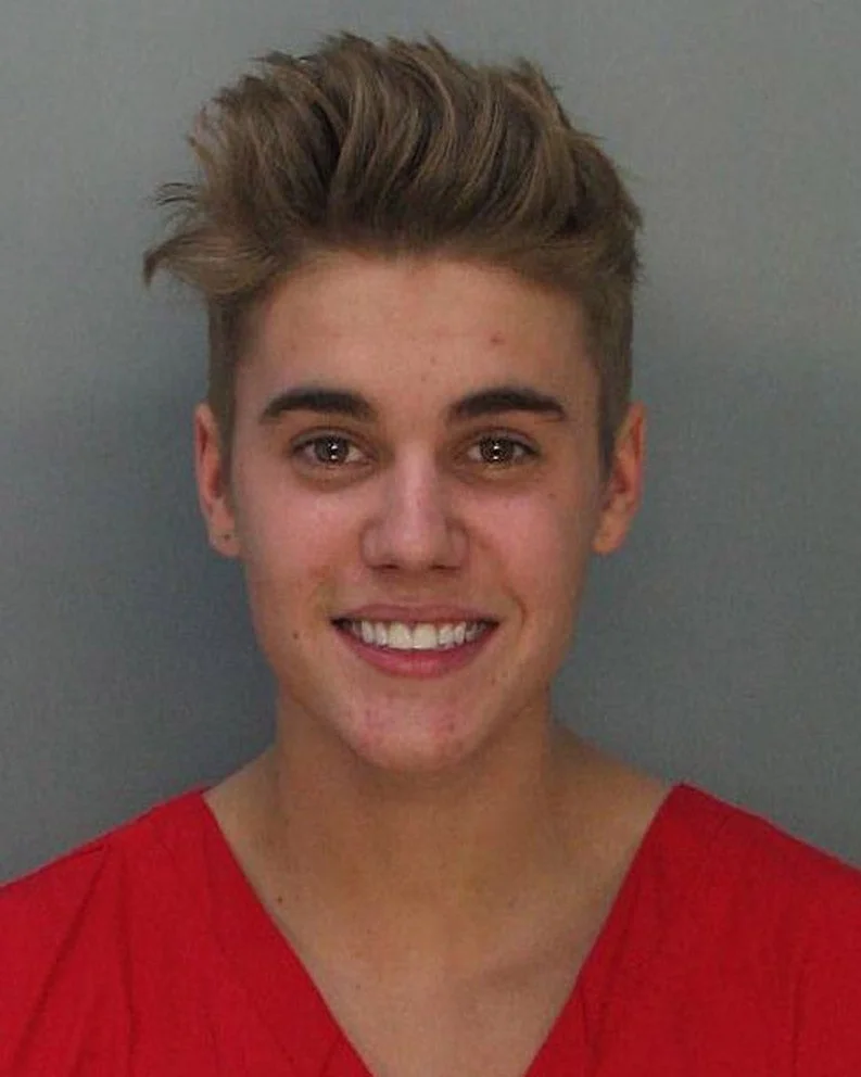 A person with short styled hair is wearing a plain shirt and smiling at the camera, against a neutral background