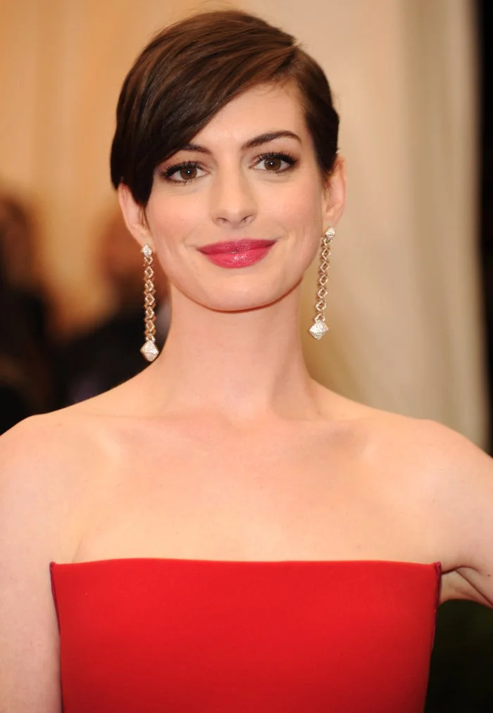 Person in elegant strapless gown with drop earrings, short hairstyle, and natural makeup poses on a formal event backdrop