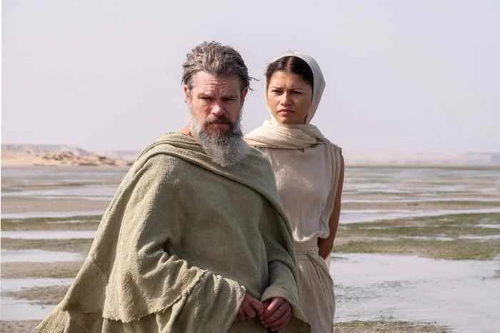 Two actors in desert costumes stand in a barren landscape, one with a beard and cloak, the other with a headscarf and sleeveless robe