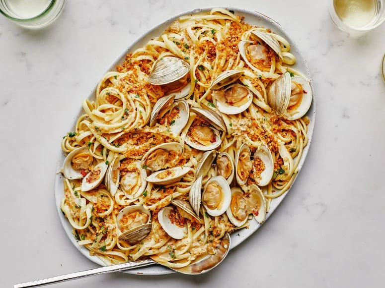 A large platter of linguine with clams alongside a bottle of white wine and two glasses.