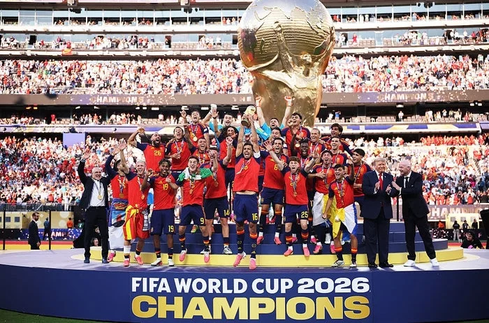 Spanish team celebrating FIFA World Cup 2026 win on stage in front of a large trophy, holding flags and wearing team uniforms, surrounded by a cheering crowd