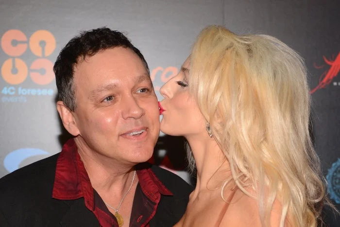A woman with long hair kisses a smiling man on the cheek at an event
