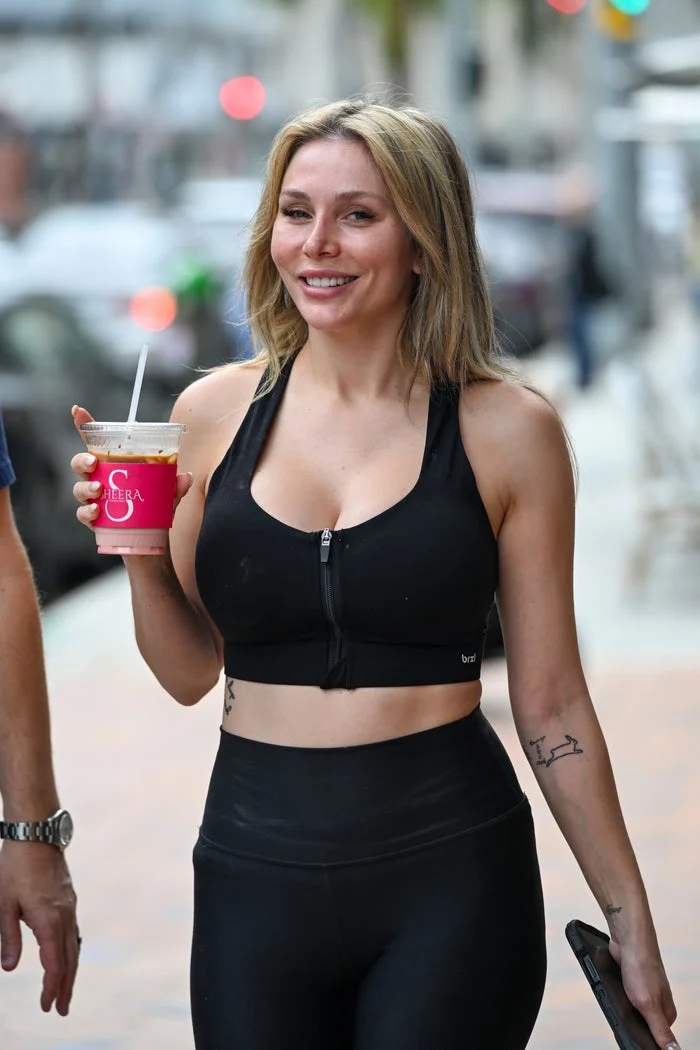 Person in a zipped sports bra and leggings holding a drink on a city sidewalk, smiling casually