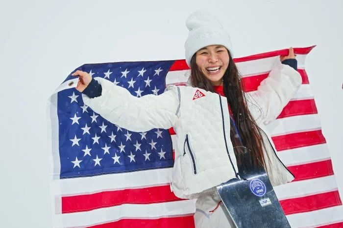 Person holding a U.S. flag and wearing a white jacket and beanie, smiling excitedly while standing with a snowboard