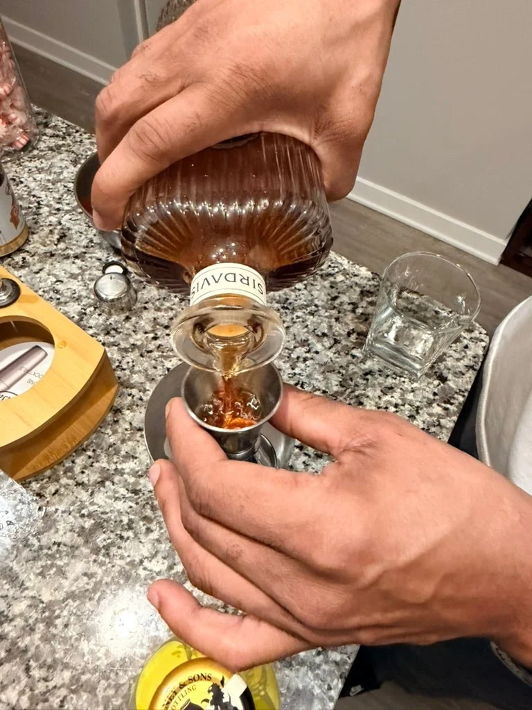 Person pouring brown liquor from a bottle into a jigger on a kitchen counter with glassware and a wooden container nearby