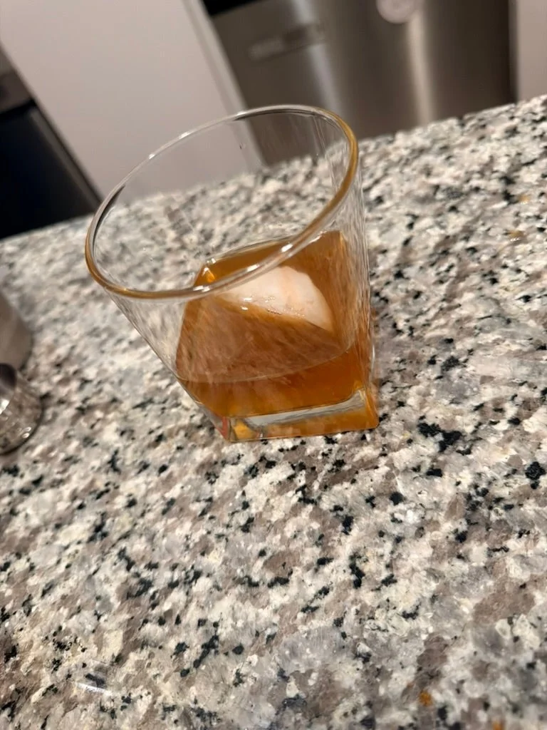 A glass of whiskey with a large ice cube on a granite countertop