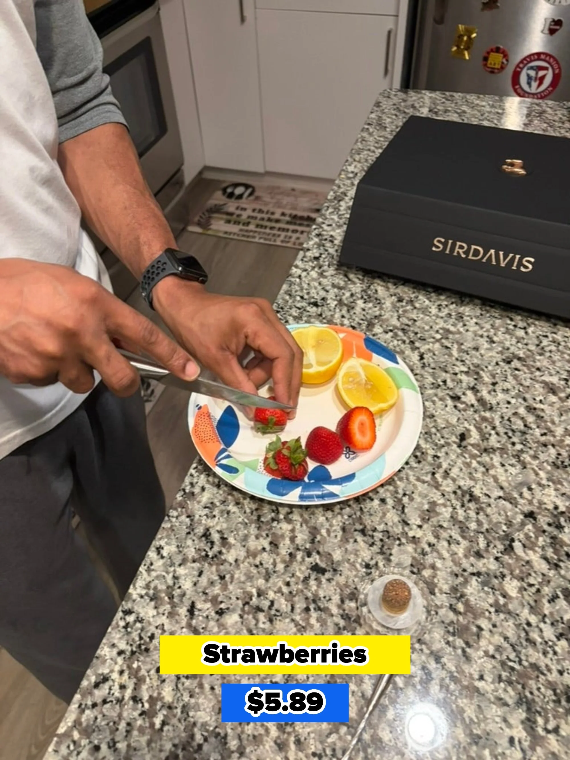 A person prepares strawberries and lemon slices on a plate in a kitchen with a box labeled "SIRDAVIS" nearby