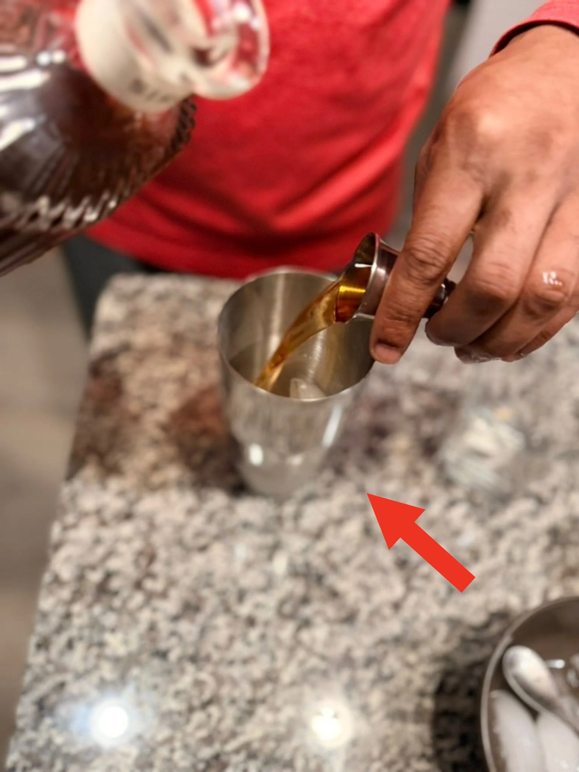 Person pouring a dark liquid from a jigger into a metal shaker on a granite countertop