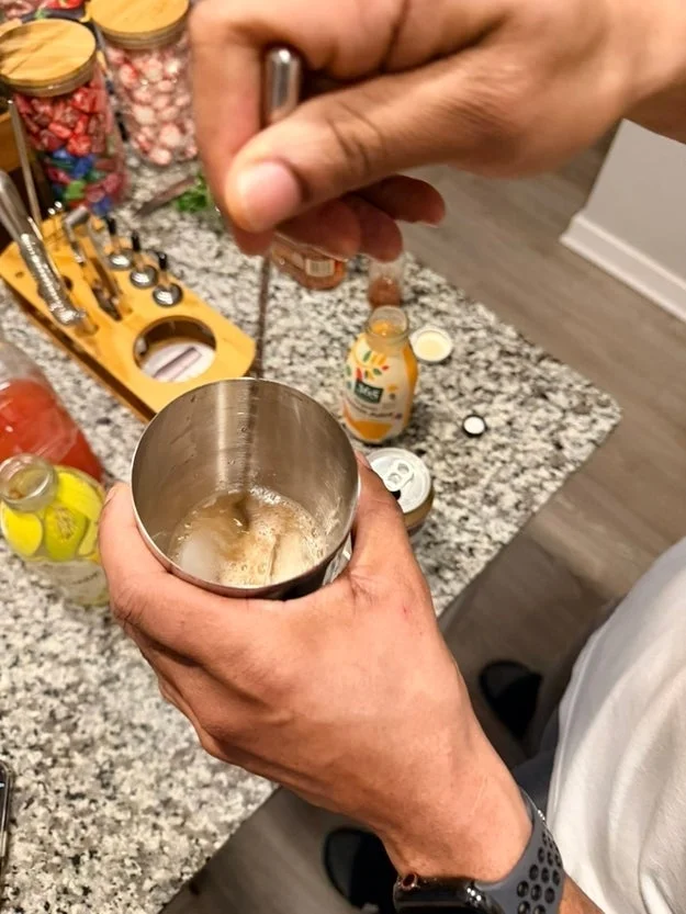 Person mixing a drink with a stirrer in a metal cup on a countertop with various drink ingredients and tools
