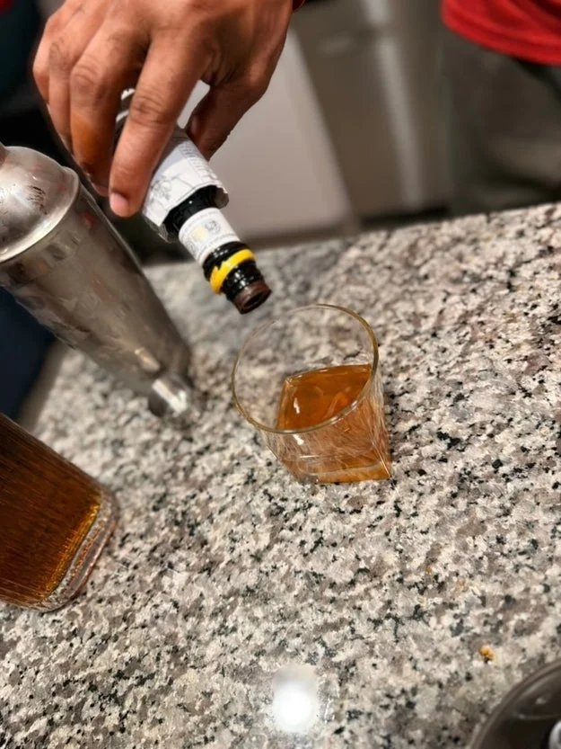 A hand pours bitters into a glass of amber liquid on a granite countertop next to a cocktail shaker