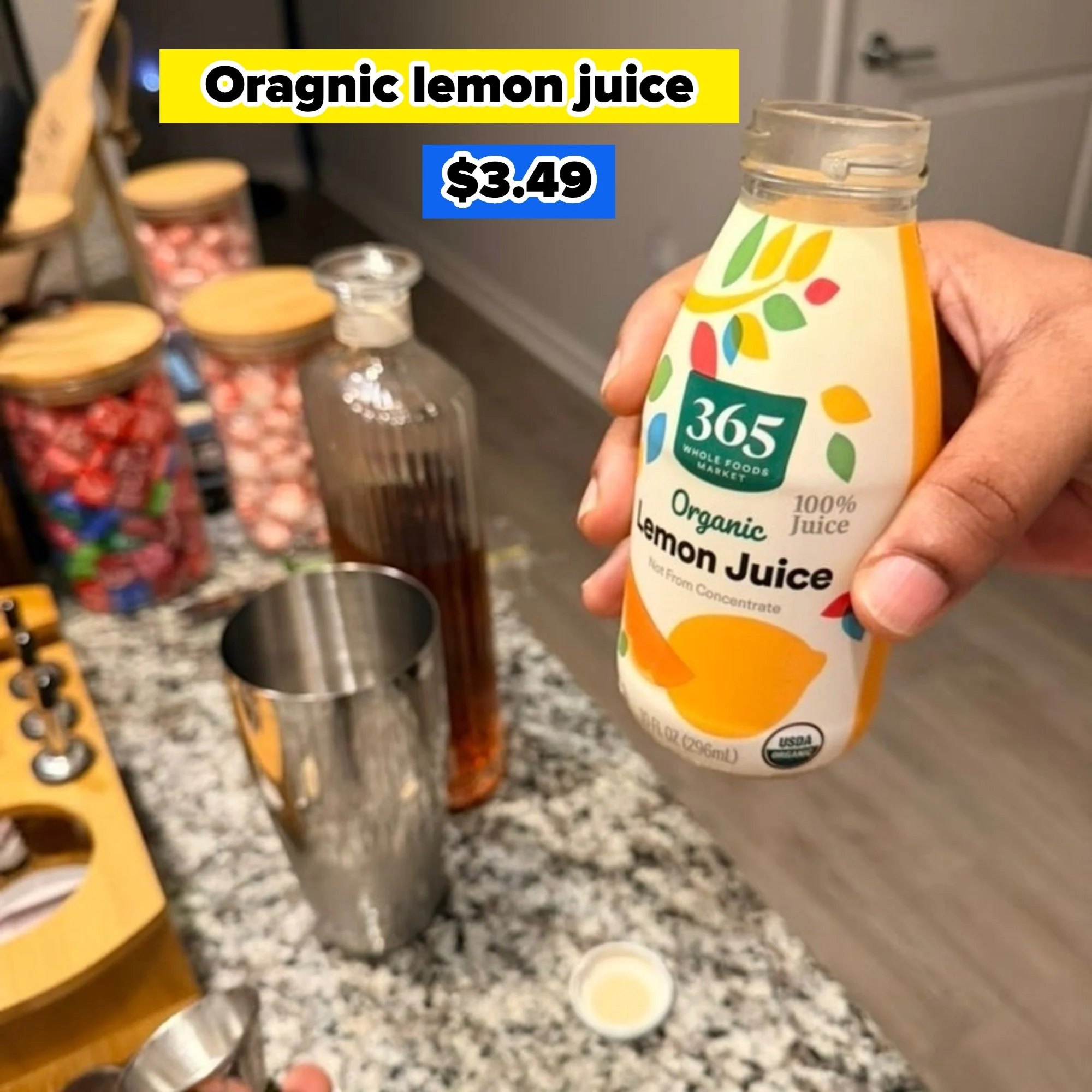 A person holds a bottle of organic lemon juice over a counter with a cocktail shaker, a liquor bottle, and candy jars nearby