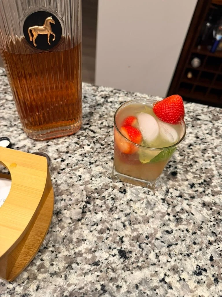 Glass of cocktail with ice, strawberries, and lime on a granite countertop next to a bottle of liquor with a horse logo