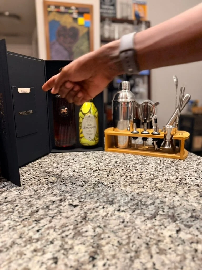 A hand reaches for a cocktail kit on a countertop, featuring spirits, mixers, a shaker, and bar tools neatly organized
