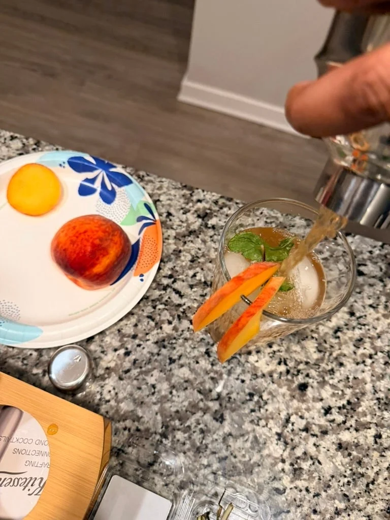 A hand pours a cocktail into a glass with ice, garnished with mint and peach slices. Nearby, a plate holds a halved peach