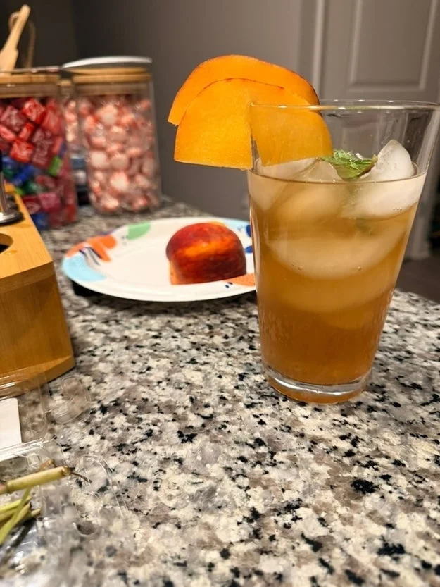 A refreshing cocktail in a tall glass with ice, garnished with peach slices, sits on a speckled countertop with snacks in the background
