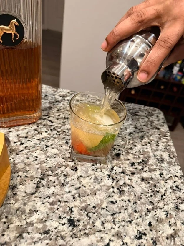 A hand pours a cocktail from a shaker into a glass with ice, mint, and fruit on a granite countertop. A whiskey bottle is nearby