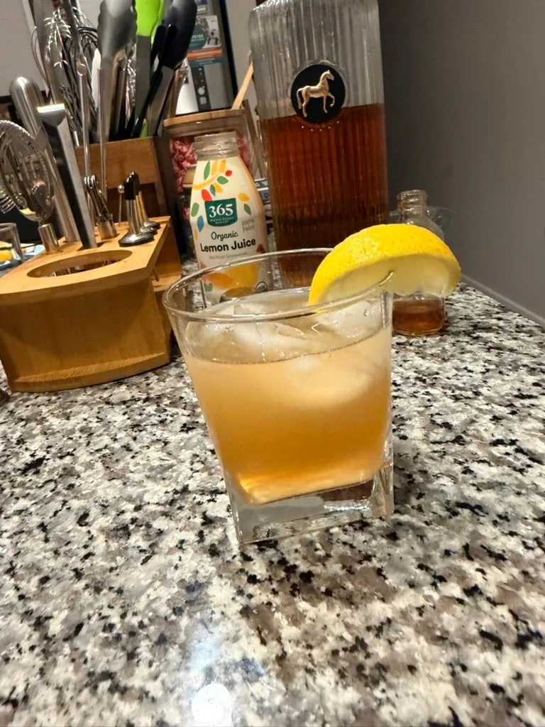 A glass of iced tea cocktail with a lemon wedge on a kitchen counter, surrounded by a knife block and a bottle labeled "Organic Lemon Juice."