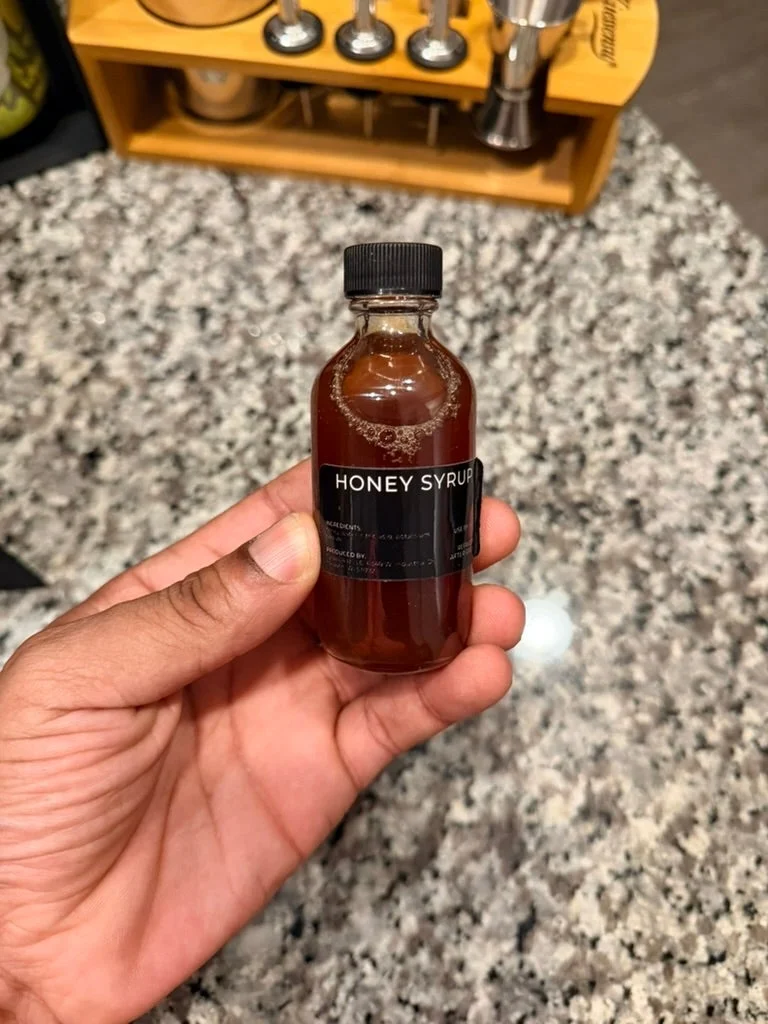 Hand holding a small bottle of honey syrup on a speckled countertop, likely used for culinary or cocktail purposes
