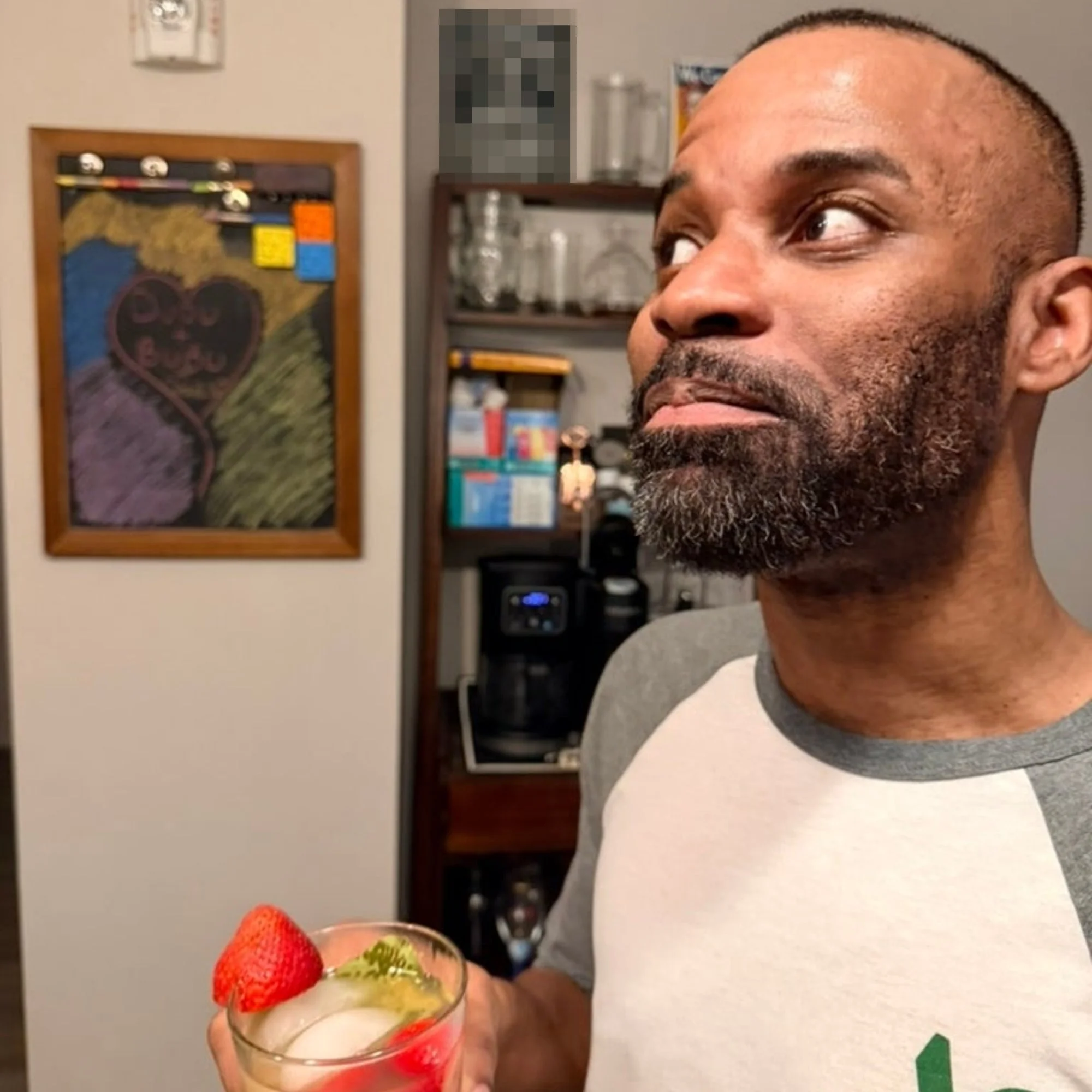 Man enjoying a beverage garnished with strawberries and herbs, standing in a kitchen. A colorful chalkboard is visible in the background