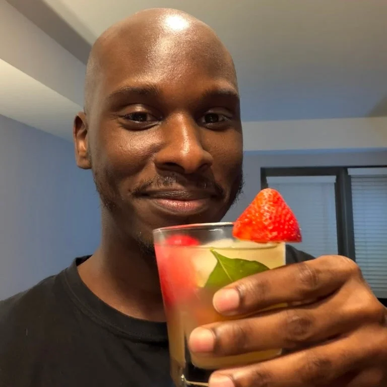Person smiling and holding a cocktail garnished with a strawberry and a green leaf