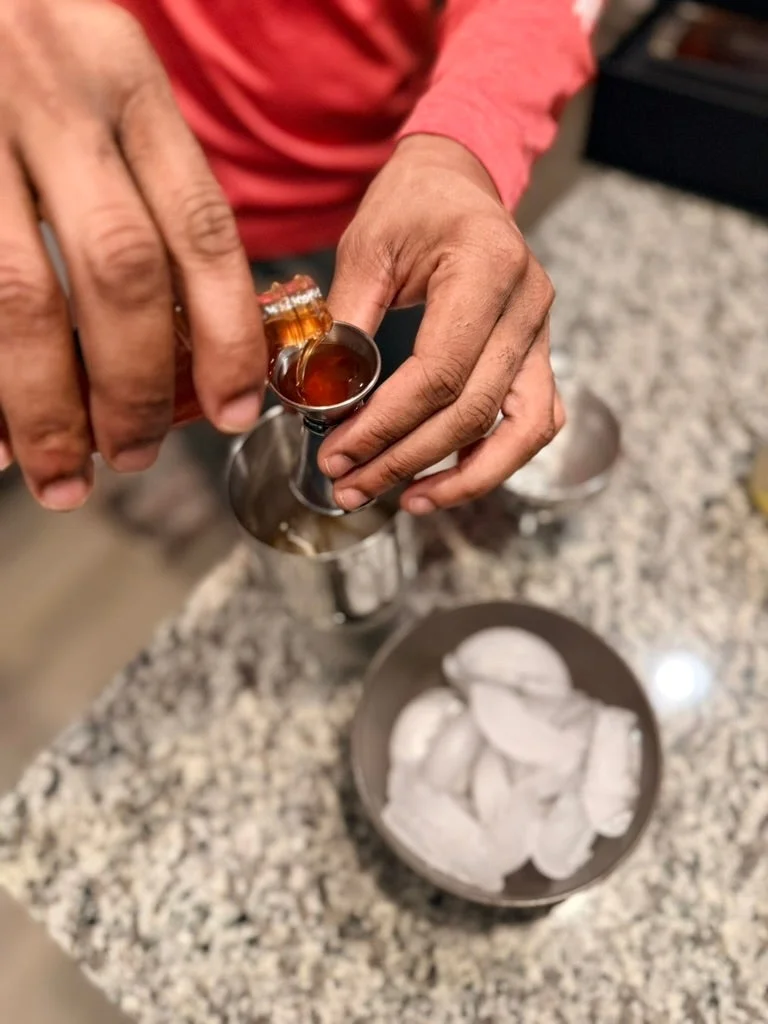 Person pouring liquid into a jigger above a bowl of ice on a granite countertop, preparing a drink