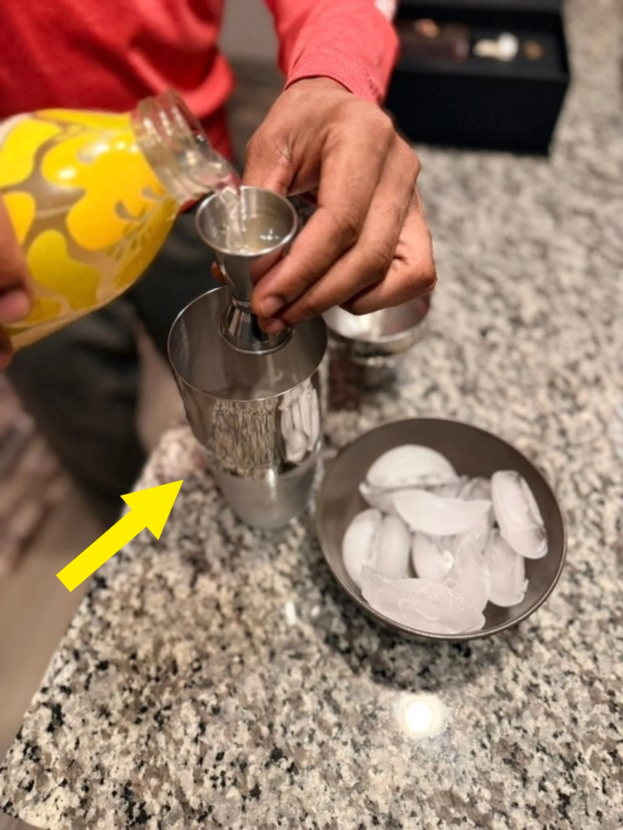 Person pouring liquid from a lemon-patterned bottle into a metal shaker, with a bowl of ice on a granite countertop