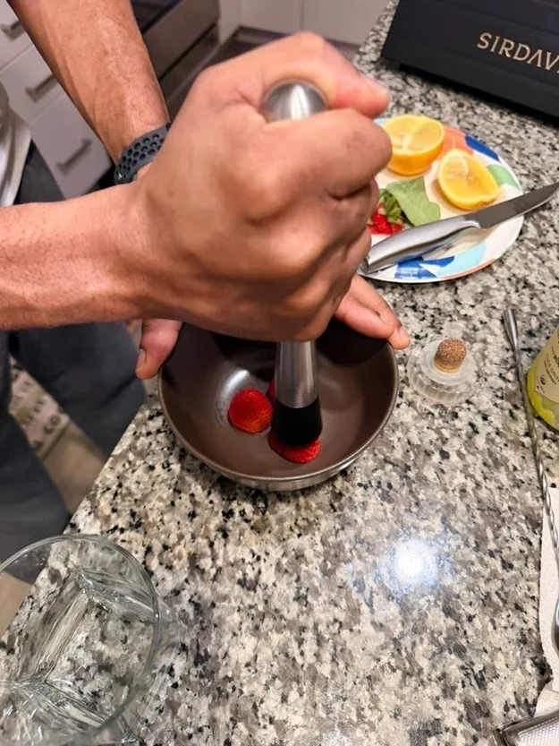 Hands mashing strawberries in a bowl on a kitchen counter, with sliced citrus and mint leaves nearby for a cocktail preparation