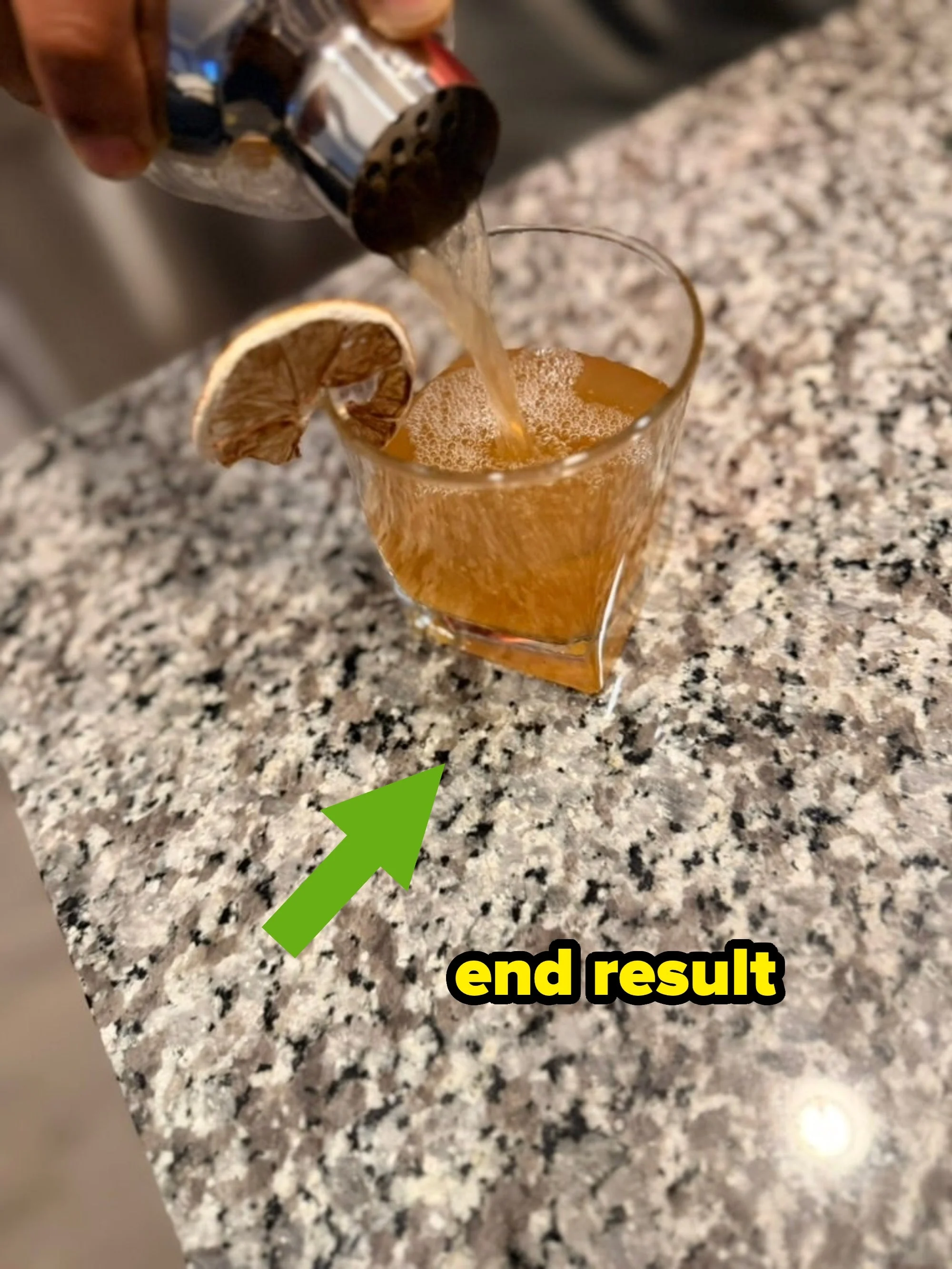 A drink is being poured into a glass garnished with a dried citrus slice on a granite countertop