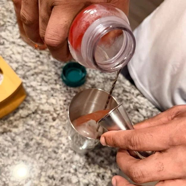 Person pouring liquid from a bottle into a measuring cup with cocoa powder on a kitchen counter
