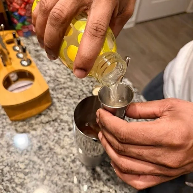 Person pouring lemon-flavored beverage into a jigger on a granite countertop with a cocktail set nearby