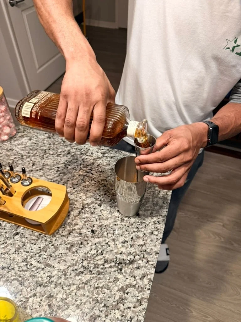 Person pouring whiskey into a cocktail shaker on a kitchen counter