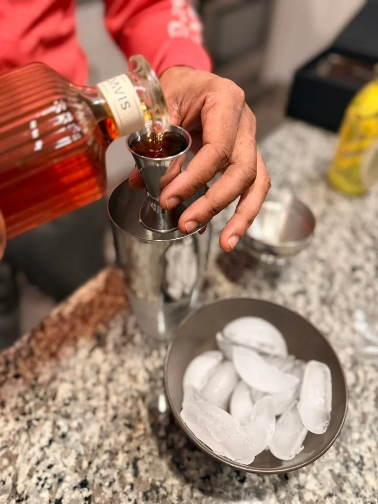 A person pours liquor into a jigger over a cocktail shaker, with a bowl of ice nearby on a granite countertop