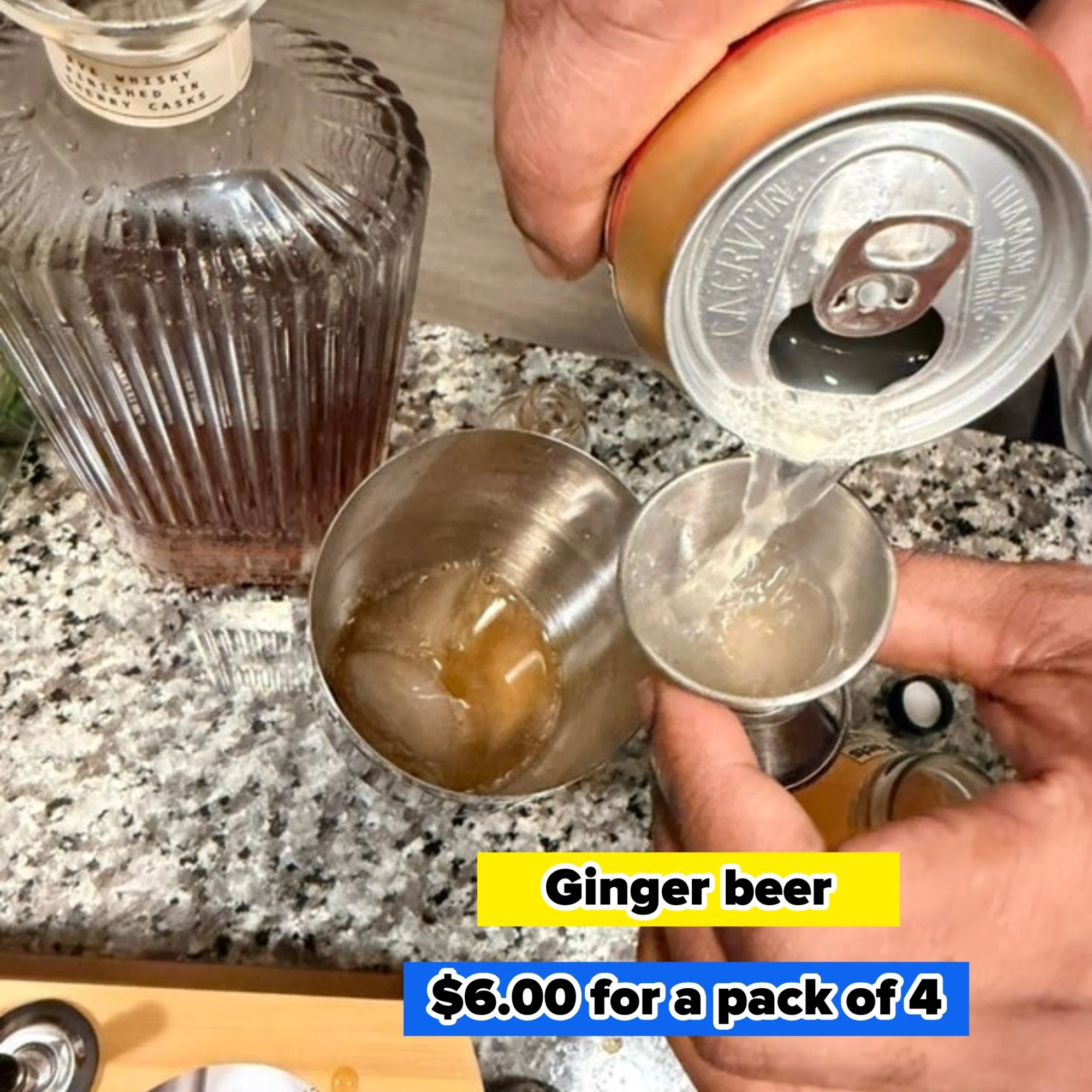 Person preparing a cocktail, pouring soda into a metal cup beside a whisky decanter on a counter