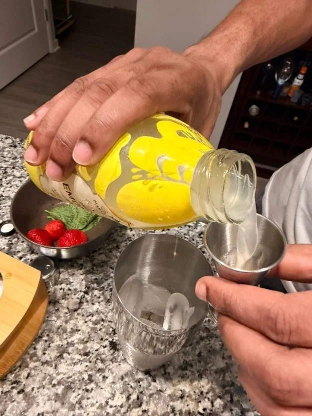 Pouring juice into a jigger over ice in a glass, with strawberries and limes nearby on a countertop