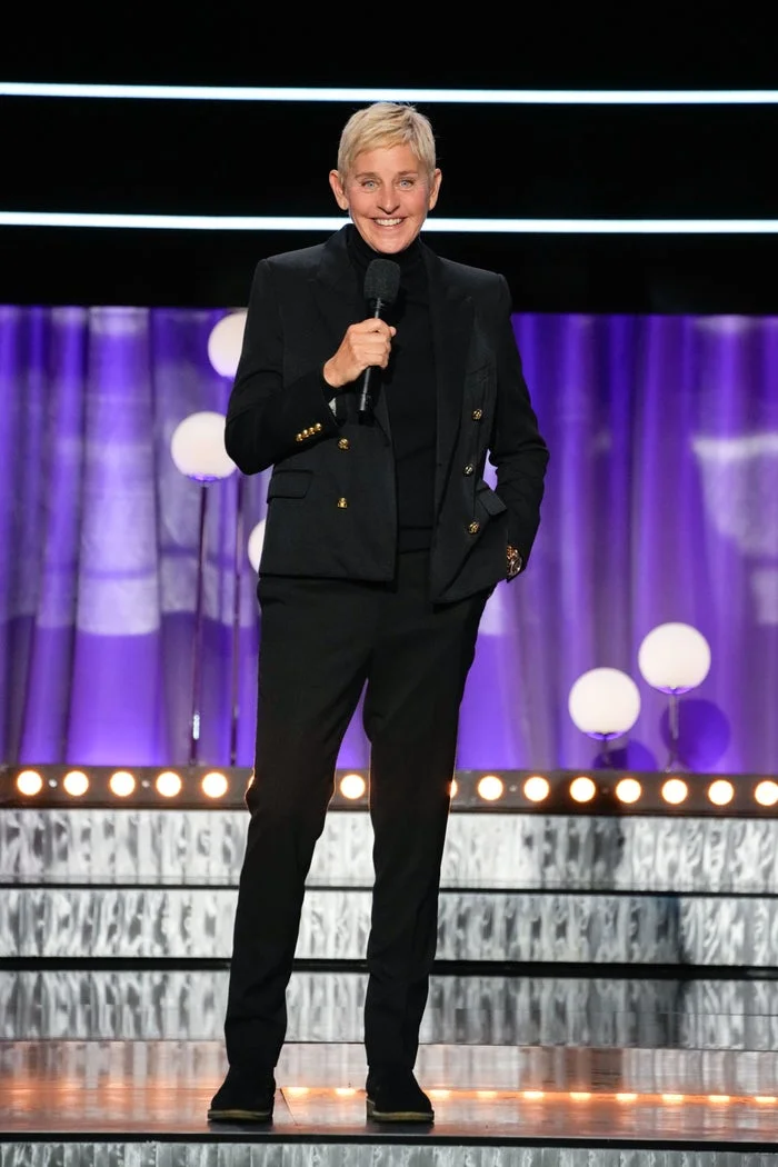Ellen DeGeneres in a black suit holding a microphone, standing on a stage with purple curtains and round lights in the background