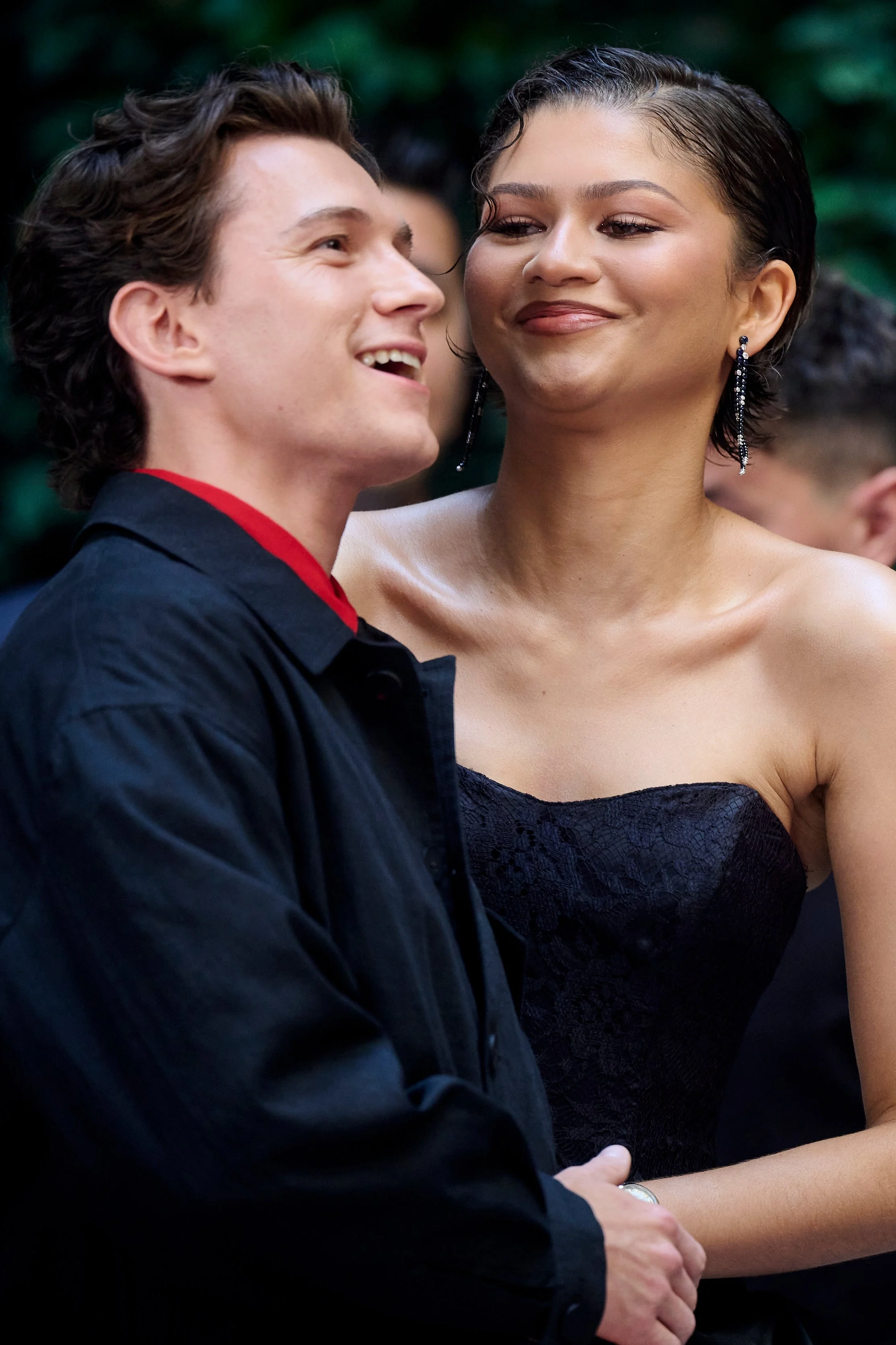 Two people smiling, one in a dark jacket with a red shirt, the other in a strapless dress with earrings